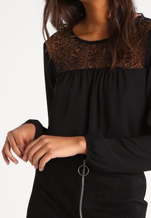 Femme portant un haut noir à manches longues avec un détail en dentelle à l’encolure et une fermeture éclair avec un tirette rond sur une jupe ou un pantalon noir.