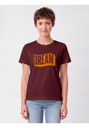 Jonge persoon met kort haar die een bruin t-shirt draagt met het woord "DREAM" in grote oranje letters, gecombineerd met lichtblauwe spijkerbroek.