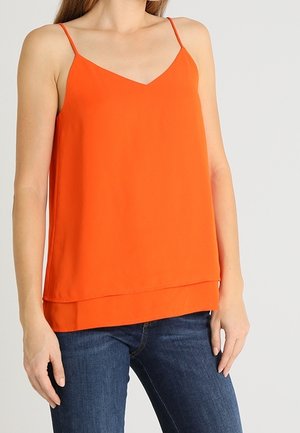 Kobieta ubrana w warstwową pomarańczową bluzkę na ramiączkach i niebieskie dżinsy, stojąca na tle jednolitego jasnego tła.