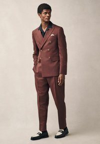 Traje de corte cruzado en tejido burdeos oscuro con detalles en negro, combinado con zapatos en blanco y negro. Destaca el detalle del pañuelo en el bolsillo y el ajuste slim.