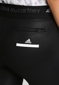 Zbliżenie czarnych legginsów sportowych z logo Adidas i Stella McCartney, z małą zapinaną na zamek kieszenią z tyłu oraz białą bluzką z siateczki.