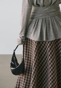 Graues langärmeliges Oberteil mit gerafftem Rückendetail, kombiniert mit einem braun karierten Rock. Eine kleine schwarze Handtasche wird in der Hand gehalten.