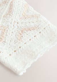 Valkoinen virkattu viltti, jossa on teksturoidut, kohotetut kuviot ja vaaleanbeiget yksityiskohdat. Viimeistelty sahalaitaisella reunuksella ja rei'itetyillä yksityiskohdilla.