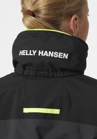 Černá nepromokavá bunda s vysokým límcem, na zadní straně s bílým logem "HELLEY HANSEN" a reflexní žlutou akcentní stužkou.