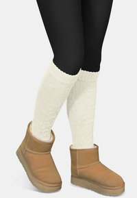 Braune Wildleder-Stiefeletten mit weichem Innenfutter, kombiniert mit weißen, kabelgestrickten Stulpen und schwarzen Leggings, die einen gemütlichen Winterlook zeigen.