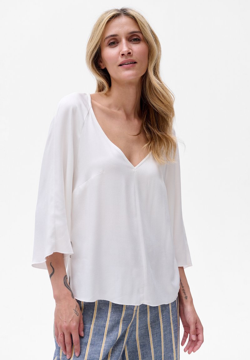 Weiße, leichte Bluse mit V-Ausschnitt, 3/4-Ärmeln und lockerer Passform; glatte Textur mit dezentem Drapierungsdetail. Kombiniert mit gestreiften Hosen.