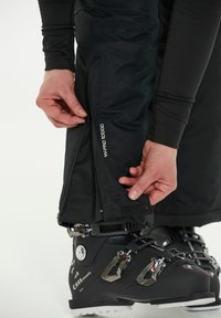 Mustat hiihtohousut, joissa on vedenpitävä vetoketju, säädettävä hihansuu ja tarrakiinnitys, logoprintillä varustettuna. Hiihtosukan osa näkyy osittain.