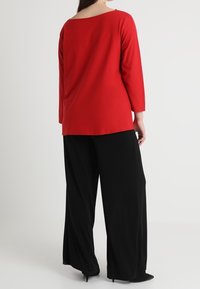 Maglione rosso a maniche lunghe con collo a barca ampio, abbinato a pantaloni neri larghi e tacchi, che evidenziano una texture del tessuto morbida.
