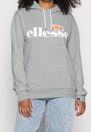 Henkilö, jolla on vaaleanharmaa huppari, jossa valkoiset nyörit ja valkoinen "ellesse"-logo oranssilla puolikuutiolla, yhdistettynä vaaleansiniin farkkuihin.