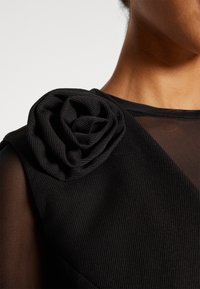 Černý oděv s texturovaným tělem, kontrastním průhledným rukávem a výraznou černou látkovou růží na rameni.