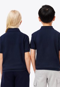 Kaksi lasta seisoo vierekkäin selkä kameraan päin, pukeutuneina tummansinisiin polo-paitoihin, yhdellä tummat shortsit ja toisella vaaleanharmaat cargo-shortsit.