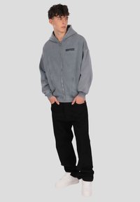 Siva jopa z zadrgo z logotipom, ima sproščen kroj, kombinirana s črnimi hlačami in belimi supergami. Gladek material in minimalni detajli.