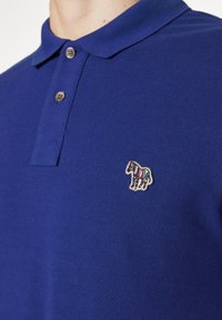 Polo bleu avec deux boutons et un petit logo zèbre multicolore brodé sur la poitrine gauche.