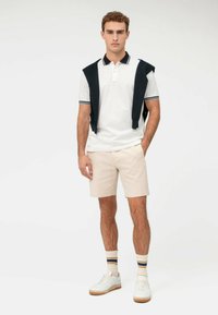 Weißes Poloshirt mit marineblauem Kragen, beiges Shorts und weiße Sneakers. Gestrickter Pullover über die Schultern drapiert; gestreifte Socken sorgen für das gewisse Etwas.
