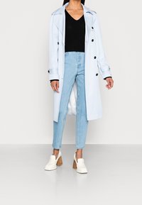Abrigo trench de color azul claro con botones negros, camiseta de cuello en V de canalé negro, jeans de tiro alto azul claro y zapatos de tacón bloque blancos.