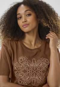 T-shirt en coton marron avec un motif brodé crémeux décoratif au centre, un col rond et des manches courtes.