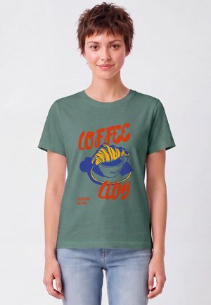 Jeune femme portant un t-shirt vert avec un texte rouge "Coffee Club" et un graphisme d'une tasse avec un croissant, associé à un jean bleu clair.