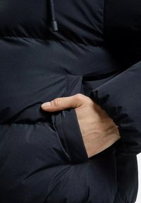 Granatowa kurtka puchowa z ręką w bocznej kieszeni. Materiał jest gładki, z pikowanym wykończeniem, z regulowanym ściągaczem przy kołnierzu.