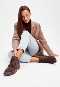 Bottines en suede marron avec un laçage, semelles en caoutchouc, bouts ronds et talons plats. Portées avec un jean bleu clair et un blazer beige.