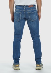 Blå denimjeans med slim fit, med två bakfickor och en brun läderlapp vid midjan, texturerad yta och ljus blekning.