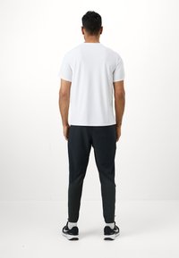 Weißes T-Shirt mit kurzen Ärmeln und schwarze, schmal geschnittene Hose mit strukturierten Seitenpanelen, getragen mit schwarzen Sportschuhen und weißen Socken.