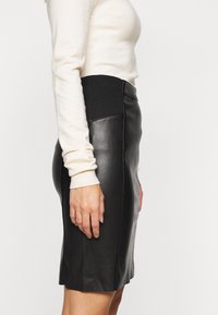 Femme portant une jupe crayon en cuir noir avec une taille élastique haute et un haut blanc moulant à manches longues, vue de profil.