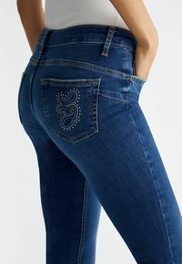 Jeans in denim blu scuro con vestibilità slim, vita media e cuciture decorative con strass sulla tasca posteriore.
