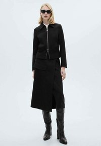 Giacca nera con zip frontale dal design strutturato, abbinata a una gonna nera avvolgente con tasca frontale. Il modello indossa stivali neri al ginocchio e occhiali da sole.