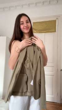 Νεαρή γυναίκα με μακριά καστανά μαλλιά κρατάει ένα μπεζ σακάκι σε ένα λευκό δωμάτιο με κλειστές διπλές πόρτες στο φόντο.