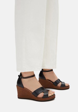 Czarne skórzane sandały na koturnie z grubym korkowym obcasem, otwartym noskiem i paskiem wokół kostki. Połączone z białymi spodniami o szerokich nogawkach.