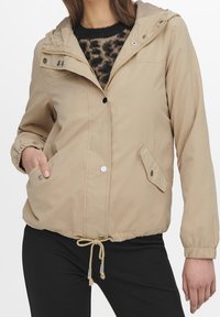 Beige hooded jacket with buttons and zipper, elastic cuffs, drawstring waist, worn over leopard print sweater and black pants.