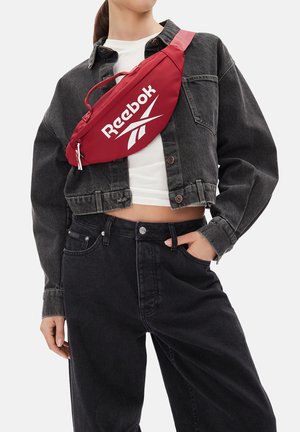 Kobieta w czarnej, krótkiej kurtce dżinsowej, białym topie odsłaniającym brzuch, czarnych dżinsach oraz czerwonej torebce Reebok przewieszonej przez pierś.