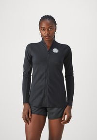 Black long-sleeve zip-up top with ribbed texture and a round logo accent on the chest, paired with dark shorts.