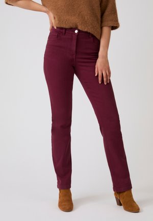 Donna che indossa jeans bordeaux a gamba dritta, maglione marrone peloso e stivaletti in camoscio color beige, in piedi con una mano sul fianco.