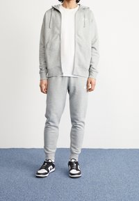Ljusgrå zip-up hoodie med dragsnören och fickor, kombinerat med matchande grå sweatpants. Svart och vita sneakers fulländar outfiten.
