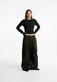 Schwarzes, geripptes Langarmshirt kombiniert mit olivgrünen, weit geschnittenen Hosen mit elastischem Bund und tiefen Seitentaschen. Glänzende schwarze Schuhe.