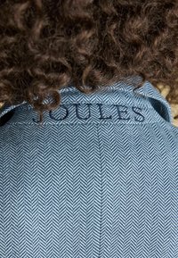 Persoană cu păr creț, purtând o jachetă albastră cu model în chevron, cu inscripția "JOULES" brodată pe guler.