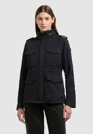 Młoda kobieta w czarnej kurtce z czterema klapkowymi kieszeniami, stojąca na tle jednolitego jasnego tła.