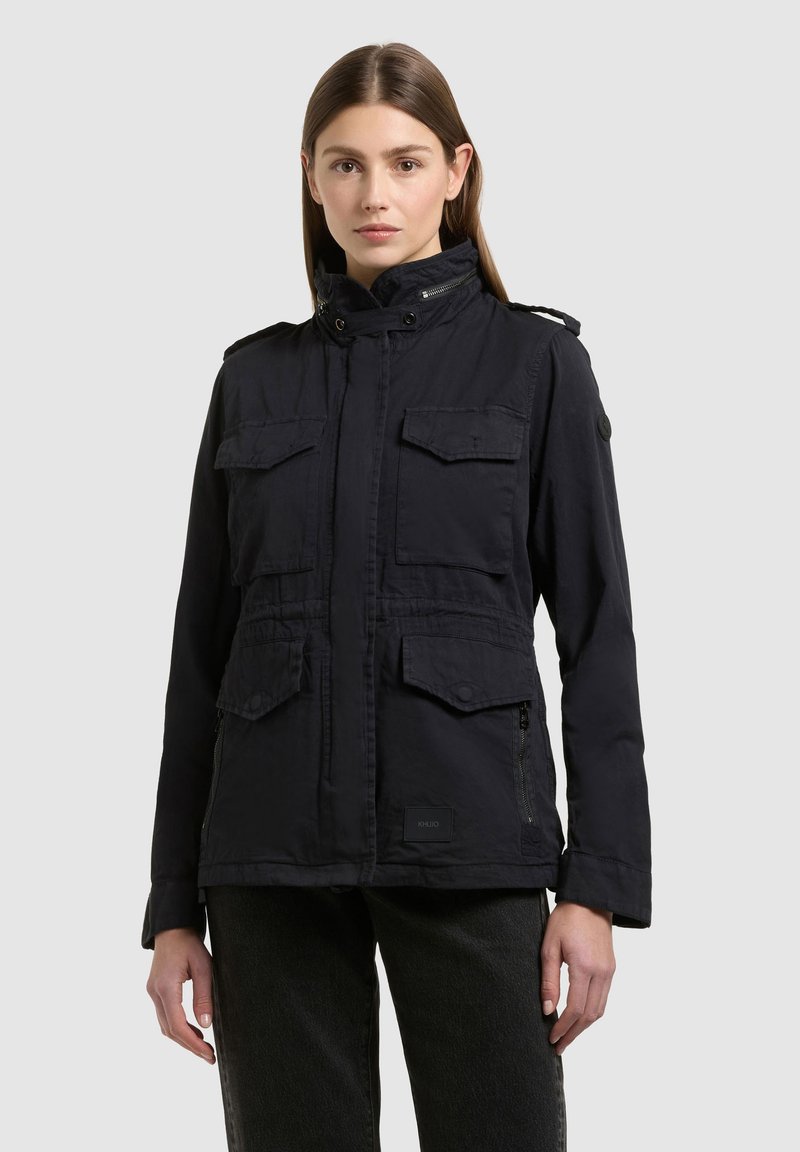 Jeune femme portant une veste noire avec quatre poches à rabat, debout devant un fond clair uni.