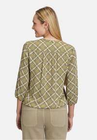 Zielona bluzka w wzory z trzema czwartymi rękawami, z geometrycznym kwiatowym wzorem w kolorze żółtym i różowym, na jasnym tle.