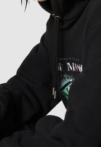 Černá mikina s kapucí, která má grafický potisk se zelenými a fialovými akcenty. Obsahuje šňůrkovou kapuci a žebrované manžety. Měkká fleecová textura.
