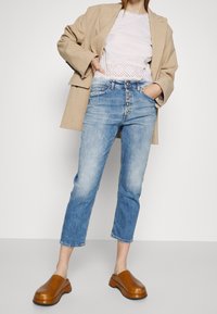 Beige oversized kavaj, vit skjorta med ögonbrynssöm, ljusblå raka jeans med knapplås och bruna plattformsträklor.