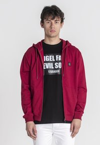 Crvena zip-up dukserica s teksturiranim finišom i prednjim džepovima, složena preko crne majice s grafikom koja ima bijeli natpis.