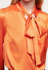 Blusa arancione in satin con collo a cravatta, dotata di dettaglio a fessura e maniche lunghe. La texture liscia e la leggera lucentezza ne esaltano l'aspetto.