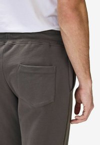 Tummanharmaat verkkarit, joissa on teksturoitu pinta, takana on paikkatasku ja joustava vyötärö. Lähikuva takanäkymästä ja käsien sijoituksesta.