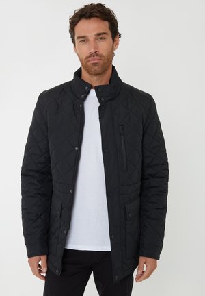 Chaqueta acolchada negra con cuello alto, bolsillo con cremallera y cierres a presión. Textura suave y ajuste entallado. Llevada sobre una camisa blanca.