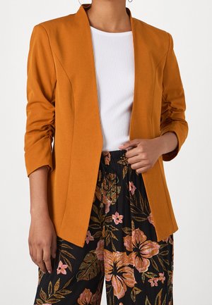 Kvinde iført en sennepsfarvet blazer over en hvid ribbet top og sorte bukser med pink og grønt blomsterprint.
