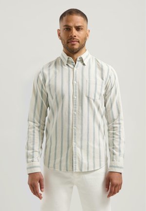 Man die rechtop staat, gekleed in een wit overhemd met verticale grijze strepen en witte broek tegen een neutrale achtergrond.