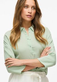 Camicia verde chiaro con maniche a tre quarti, tessuto texturizzato e bottoni abbinati. Indossata con una cintura bianca e una gonna.