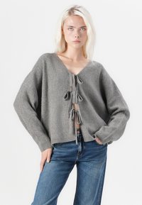 Γκρίζα πλέκτης κάρντιγκαν με V-λαιμόκοψη και εμπρόσθια κορδόνια, με μακριά μανίκια και χαλαρή εφαρμογή, συνδυασμένο με μπλε τζιν.